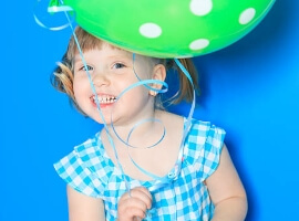 Little girl smiling and wearing a blue checkered shirt in front of a blue backdrop holding a balloon