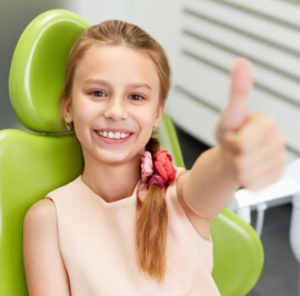 Little girl in a dentist's chair smiling and giving a thumbs up